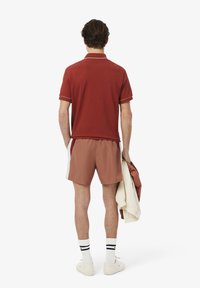 Camisa polo de manga corta color granate con ribetes blancos, combinada con pantalones cortos deportivos marrones. Modelo sosteniendo una chaqueta de color marrón claro. Calcetines blancos con rayas.