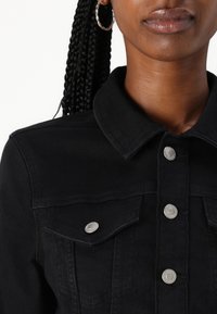 Veste en denim noir avec boutons-pression argentés, deux poches poitrine et un col classique. Équipée d'un tissu texturé et d'un design structuré.