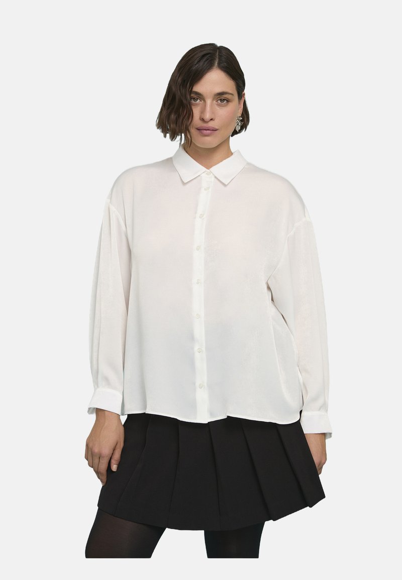 Camicia bianca con colletto, maniche lunghe e vestibilità ampia. Il tessuto sembra liscio con una leggera lucentezza. Indossata sopra una gonna nera plissettata.