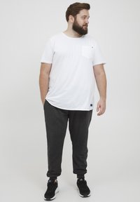 Weißes T-Shirt mit kurzen Ärmeln und einer Brusttasche, kombiniert mit dunkelgrauen Jogginghosen. Schwarze Sportschuhe mit weißen Akzenten. Neutraler Hintergrund.