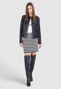 Schwarze Lederjacke, weißes T-Shirt mit Grafik, Hahnentritt-Rock, kniehohe schwarze Lederstiefel, fransiger Saum. Model steht vor einem grauen Hintergrund.