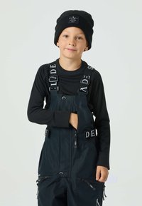 Svart ribbad långärmad tröja under mörka hängselbyxor med märkta band. Hängselbyxorna har dragkedjefickor och en framficka. Svart mössa.