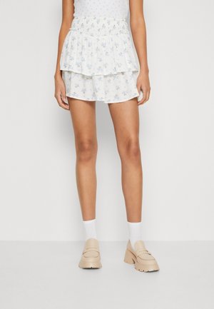 Kvinde iført hvid lagdelt mininederdel med blomster, hvide sokker og beige loafers med chunky hæl, stående mod en ensfarvet lys baggrund.
