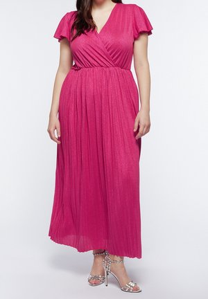 Femme portant une robe maxi plissée rose vif avec des manches volantes et des sandales argentées à talons hauts, debout devant un fond blanc.