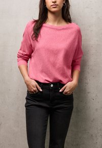 Maglione rosa con una vestibilità ampia e scollo largo, abbinato a jeans neri. Il maglione presenta polsini a costine e una texture morbida.