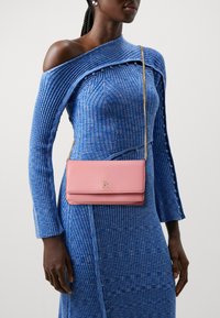 Borsa a tracolla in pelle rosa con logo dorato, abbinata a un vestito a costine blu con design off-shoulder e dettagli con bottoni.