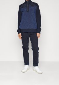 Pull-over matelassé bleu marine avec un col montant et une fermeture éclair à moitié. Manches et taille en maille côtelée. Associé à un pantalon foncé et des baskets blanches.