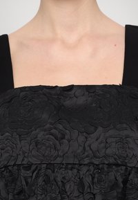 Robe noire avec un corsage texturé orné de motifs de roses brodés, décolleté carré et larges bretelles, contrastant avec une jupe lisse.