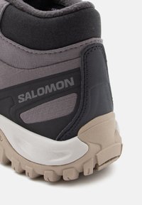 Hallhalli hallitossud, millel on tekstureeritud ülemine osa, mustad aktsendid, tugevdatud kand ja beež haardepinne. Omab "SALOMON" brändingut.