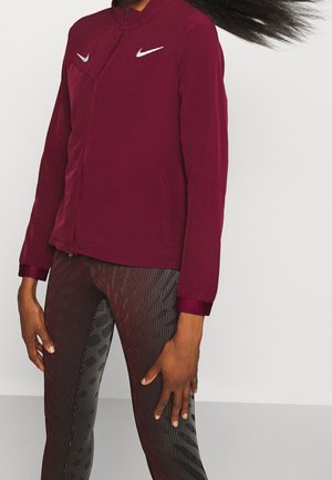 Person trägt eine burgunderrote Zip-Up-Jacke mit weißen Logos und dunkle, strukturierte Leggings, steht vor einem einfachen hellen Hintergrund.
