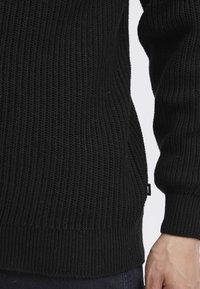 Černý pletený svetr s žebrovanou strukturou, s dlouhými rukávy a přiléhavým lemem. Na boku viditelná malá logo značka.