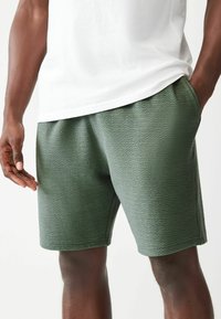 Pantaloni corti verdi con una superficie testurizzata, elastico in vita e tasche laterali, abbinati a una maglietta bianca. Dettagli del tessuto e cuciture visibili.