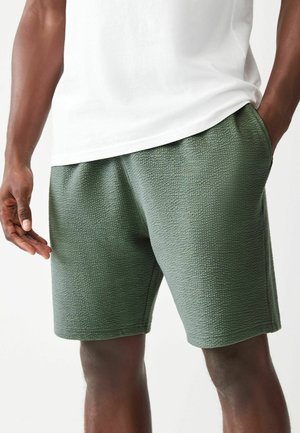 Pantaloni corti verdi con una superficie testurizzata, elastico in vita e tasche laterali, abbinati a una maglietta bianca. Dettagli del tessuto e cuciture visibili.