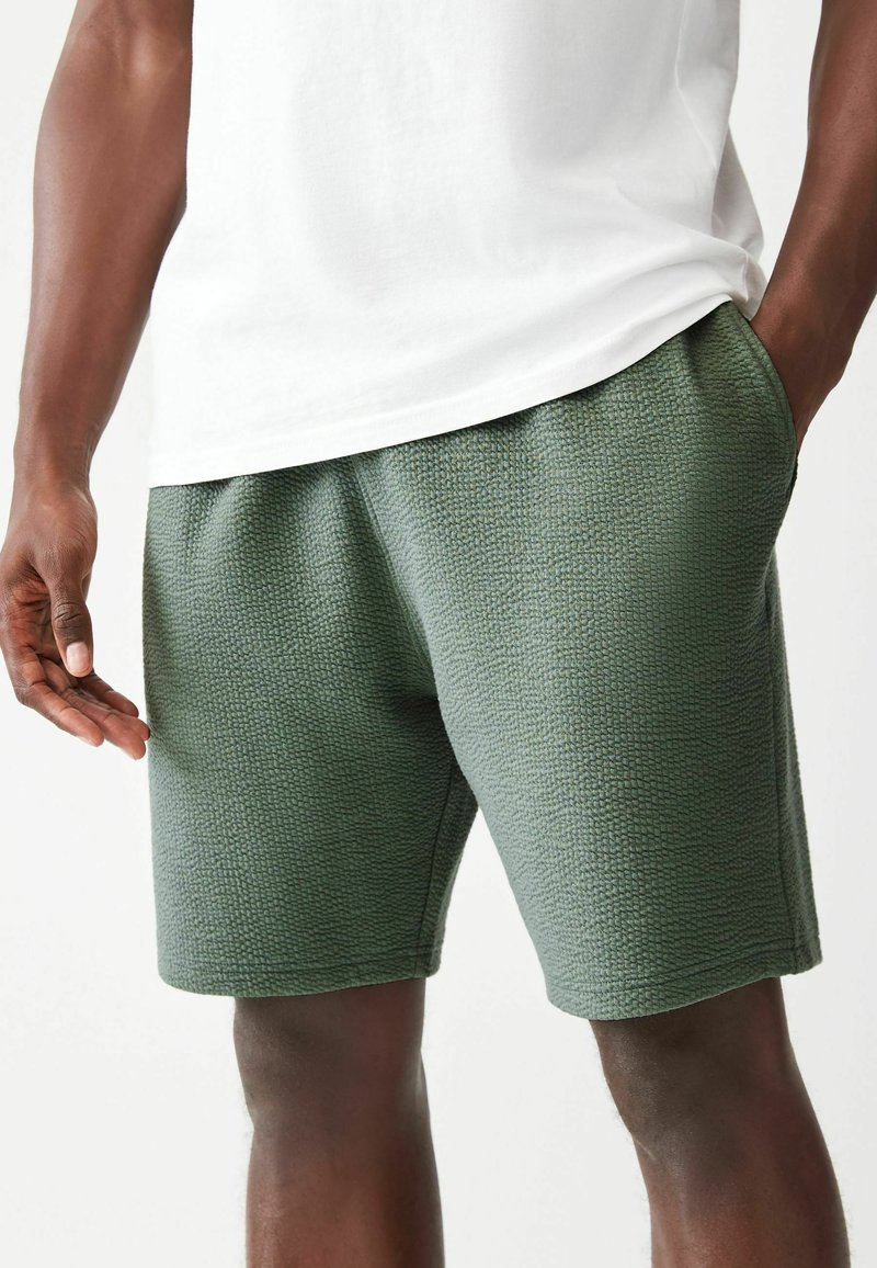 Pantaloni corti verdi con una superficie testurizzata, elastico in vita e tasche laterali, abbinati a una maglietta bianca. Dettagli del tessuto e cuciture visibili.