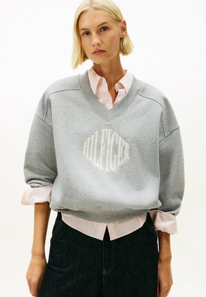 Sweat-shirt gris avec décolleté en V et logo "HILFIGER" brodé, superposé sur une chemise rayée rose et blanche. Présente une coupe ample et des poignets.