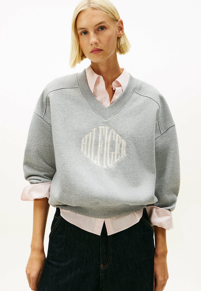 Felpa grigia con scollo a V e logo ricamato "HILFIGER", indossata sopra una camicia a righe rosa e bianche. Presenta una vestibilità ampia e polsini.
