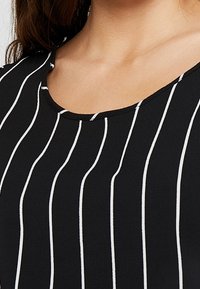 Haut en tissu noir avec des rayures verticales blanches, encolure ronde et texture lisse. La vue en gros plan met en avant le motif rayé.