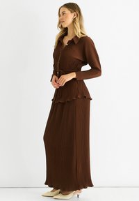 Brown pleated button-up dress with a belted waist and scalloped hem. Long sleeves with rolled cuffs, paired with pointed white heels.