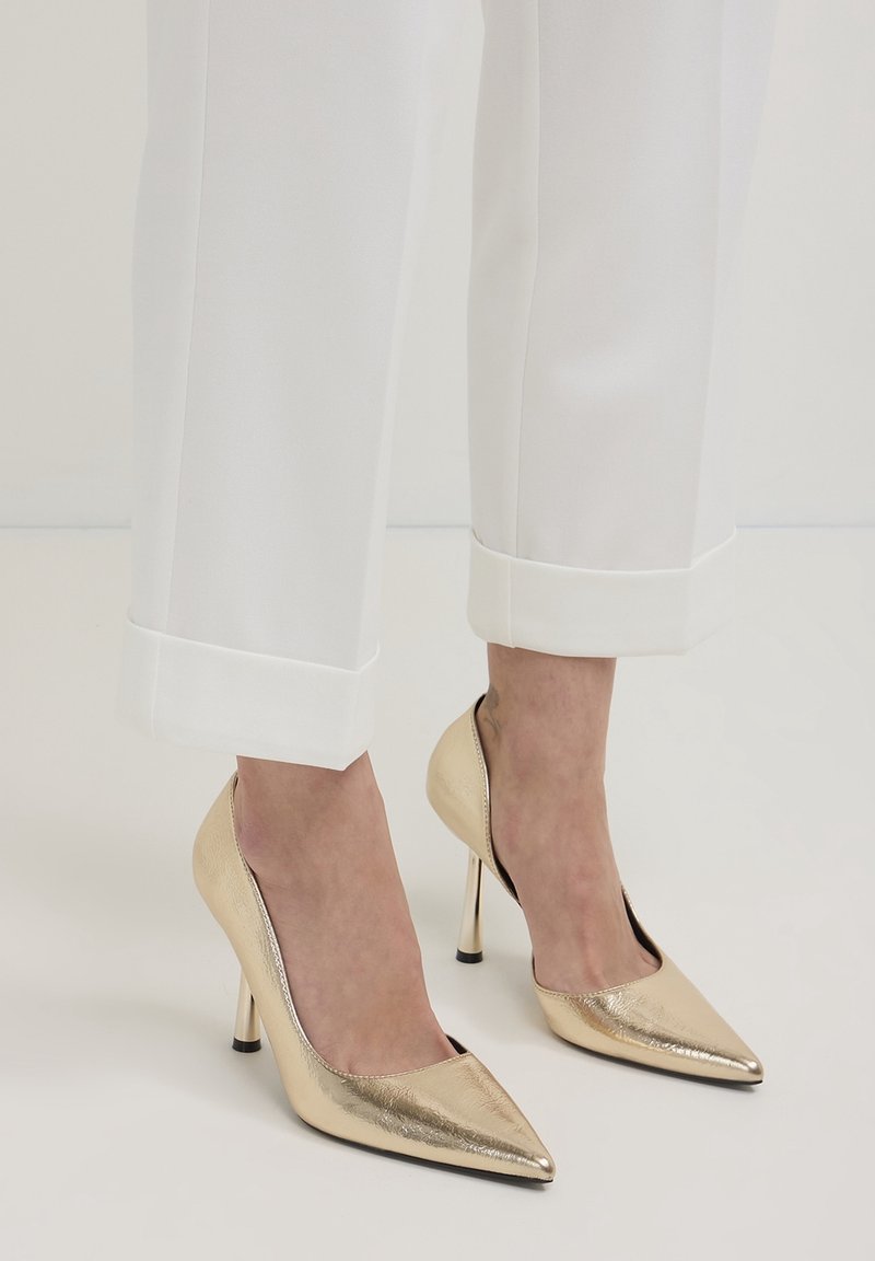 Pieds portant des escarpins à bout pointu dorés métalliques associés à un pantalon blanc court avec de larges revers.