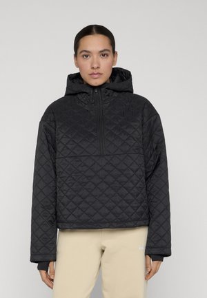 Ung kvinde iført en sort quiltet hættetrøje med lynlås foran, beige bukser, stående mod en ensfarvet lys baggrund.