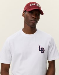 Λευκό μπλουζάκι από βαμβάκι με κοντά μανίκια και ναυτικό λογότυπο στο αριστερό στήθος. Μπορντό καπέλο με το "LES DEUX" κεντημένο στα λευκά.