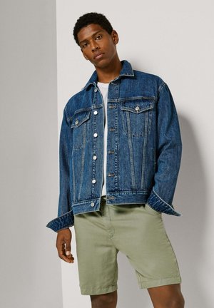 Fiatal férfi kék farmerdzsekiben, fehér pólóban és világoszöld rövidnadrágban, egy egyszerű világosszürke fal előtt áll.