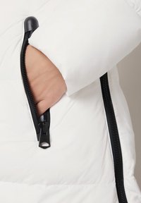 Ręka wewnątrz częściowo rozpiętej kieszeni białej kurtki puchowej z czarnymi detalami zamka.