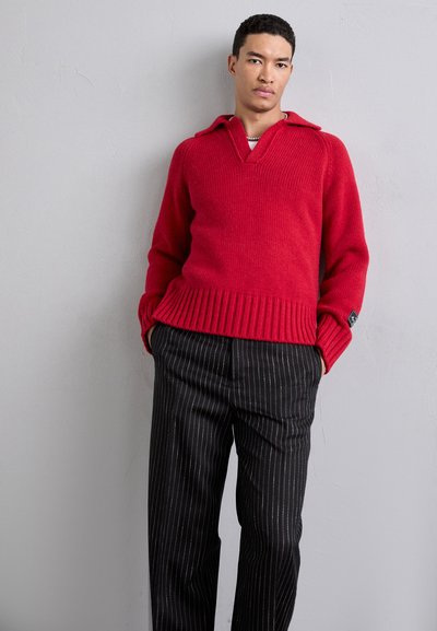 Red knit sweater with a v-neck and ribbed cuffs, paired with black pinstriped trousers. Sweater features a collar and textured fabric.