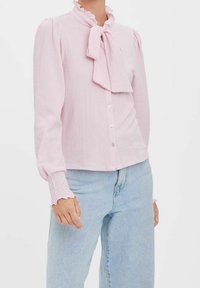 Blusa rosa chiaro con colletto a ruffle e dettaglio a fiocco. Presenta maniche lunghe con polsini arricciati e chiusura a bottoni sul davanti. Tessuto strutturato.