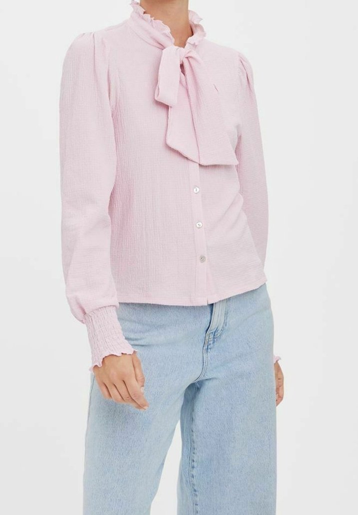 Blusa rosa chiaro con colletto a ruffle e dettaglio a fiocco. Presenta maniche lunghe con polsini arricciati e chiusura a bottoni sul davanti. Tessuto strutturato.