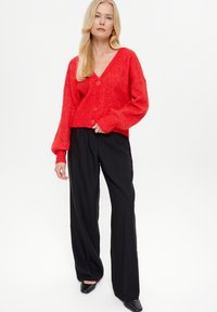 Cardigan lavorato a maglia rosso, con trama, scollo a V, maniche lunghe a sbuffo e tre bottoni davanti. Abbinato a pantaloni neri a gamba larga.