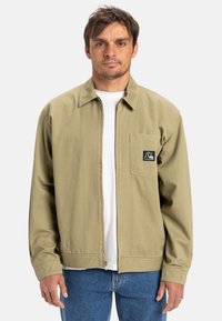 Khaki ritsjack met kraag, linkerschouderzak en logo-opdruk. Lichtgewicht stof, lange mouwen en elastische tailleband. Gedragen over een wit shirt.