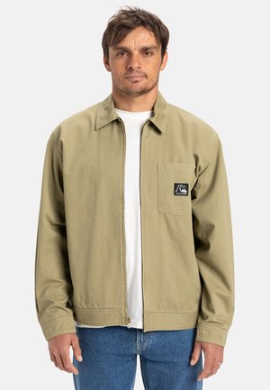 Khaki zip-up jacket with collar, left chest pocket, and logo patch. Lightweight fabric, long sleeves, and elastic waistband. Worn over a white shirt.