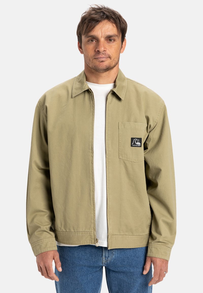 Khaki ritsjack met kraag, linkerschouderzak en logo-opdruk. Lichtgewicht stof, lange mouwen en elastische tailleband. Gedragen over een wit shirt.