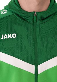 Grüne Sportjacke mit strukturiertem Muster, weißen Akzenten und einem Reißverschluss vorne. Trägt das Wort "JAKO" in fetter weißer Schrift.