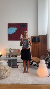 Szary top na ramiączkach trzymany w rękach, czarne szorty, stojący w nowoczesnym salonie z abstrakcyjną sztuką na ścianie, pudełkiem Zalando i różnymi meblami.