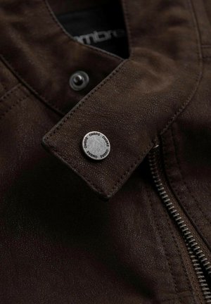 Veste en cuir marron avec une surface texturée, dotée d'un bouton-pression argenté et d'une fermeture éclair sur le devant. Le logo de la marque est visible sur le bouton.