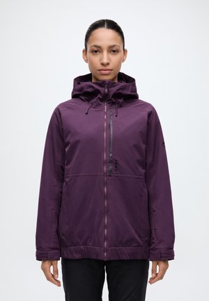 Une femme se tient face à l’objectif, vêtue d’une veste violette à capuche et à fermeture éclair ainsi que d’un pantalon noir, sur un fond blanc uni.