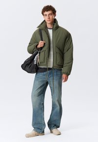 Grøn puffjakke med høj krave og to forlommer, båret over en stribet sweater. Lys blå baggy jeans og beige sko.