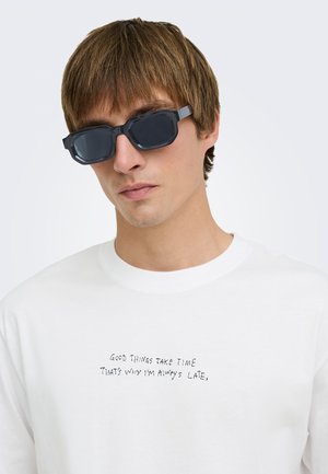 Jeune homme aux cheveux bruns portant des lunettes de soleil rectangulaires noires et une chemise blanche avec un texte indiquant "Good things take time that's why I'm always late"