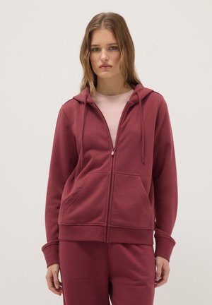 Junge Frau mit glattem Haar, die einen kastanienbraunen Reißverschluss-Hoodie und passende Hose trägt, steht vor einem schlichten hellen Hintergrund.