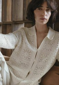 Vrouw met kort donker haar, gekleed in een crèmekleurige gehaakte cardigan met knoopsluiting, zittend tegen een houten achterwand.