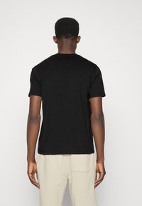 Muž stojící zády, oblečený v jednoduchém černém tričku s krátkým rukávem a světlých béžových kalhotách, na jednoduchém světlém pozadí.