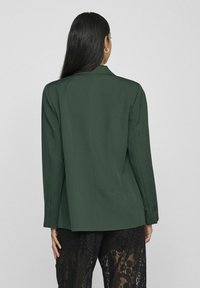 Mørkegrønn skreddersydd blazer med strukturerte skuldre og en jevn tekstur, parret med svarte blondebukser. Bakvisningen viser rene linjer og minimal detalj.