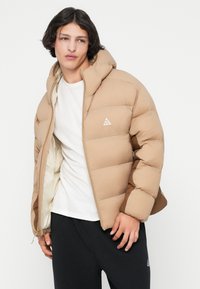 Chaqueta acolchada beige con capucha, con secciones acolchadas y un logo en el pecho. El forro interior es de color crema; diseño ligero.