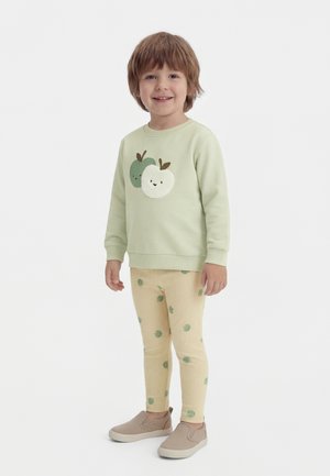 Fiatal gyermek áll és mosolyog, világos zöld pulóvert visel mosolygó almákkal, valamint bézs nadrágot zöld almamintákkal.