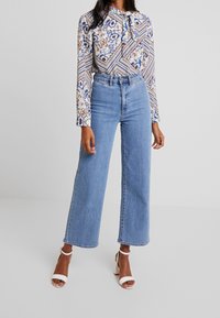 KVětovaná košile s dlouhým rukávem a vázankou na krku, kombinovaná s světlými světle modrými širokými kalhotami s vysokým pasem a bílými sandály na pásky. Hladká tkanina.