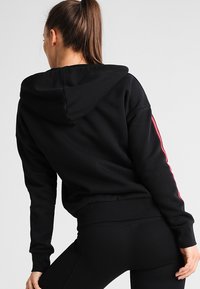 Sweat à capuche noir en tissu doux, avec une grande capuche, des manches longues et des rayures latérales roses. Taille ajustée avec des détails côtelés.