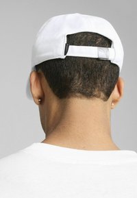 Witte honkbalpet met een contrasterende zwarte verstelbare band, voorzien van een zilveren logotag. Glad materiaal, ronde vorm, subtiele textuurdetails.