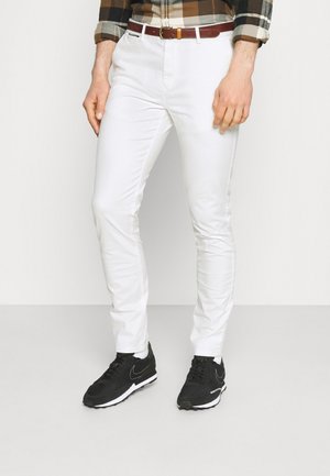 Hombre con pantalones blancos ajustados, cinturón marrón, zapatillas negras y una camisa de cuadros marrón y verde con las mangas remangadas.